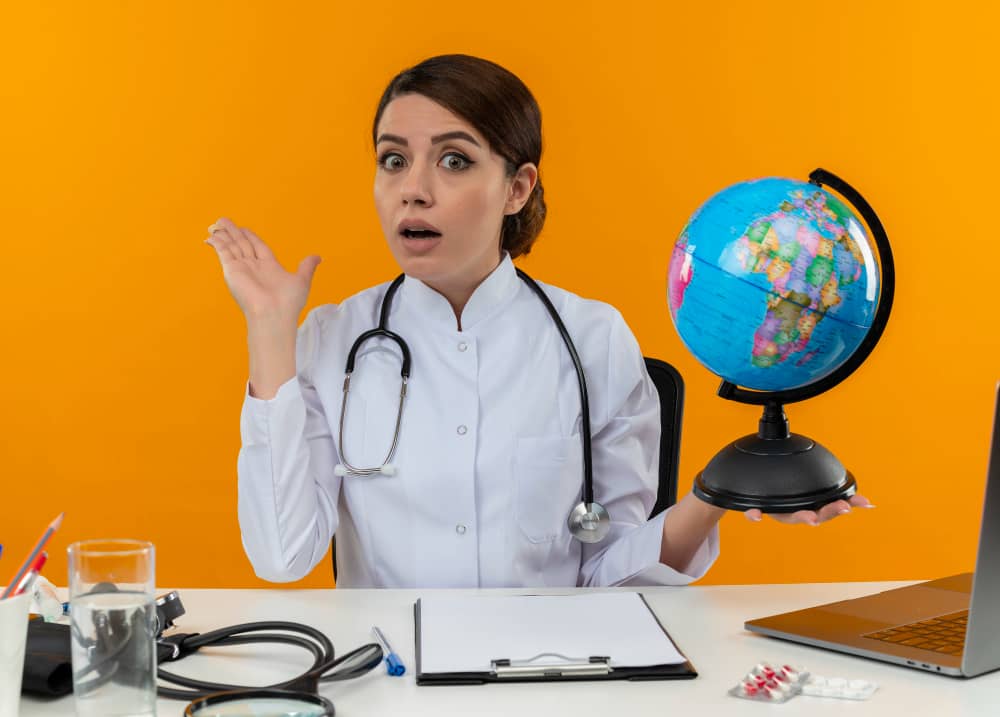 impressed-young-female-doctor-wearing-medical-robe-stethoscope-sitting-desk-with-medical-tools-laptop-holding-globe-showing-empty-hand